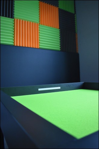 Wnętrze pokoju do gier. Stół wyłożony zielonym płótnem i zielone, porańczowe i czarne kwadraty z gąbki na ścianach.