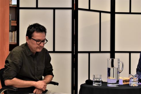Po lewej stronie siedzi mężczyzna z ciemnymi włosami, w okularach z czarnymi oprawkami, ubrany w ciemnozieloną koszulę i jasne spodnie. Po prawej stronie fragment stolika przykrytego czarnym obrusem, na którym stoją dwie szklanki i szklany dzbanek z wodą.