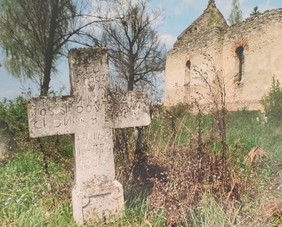 Stary kamienny krzyż wśród trawy w tle ruina kaplicy